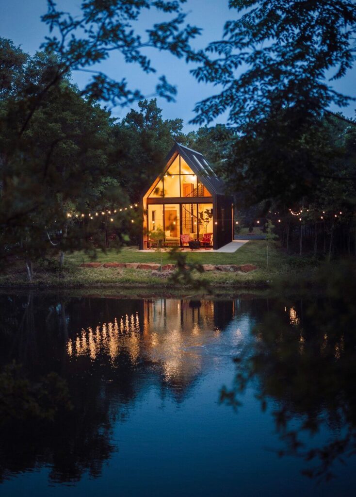 Moderna kućica s velikim staklenim prozorima i terasom. Kućica se nalazi uz jezero i okružena je drvećem. U kućici gori svjetlo, što stvara ugodnu atmosferu