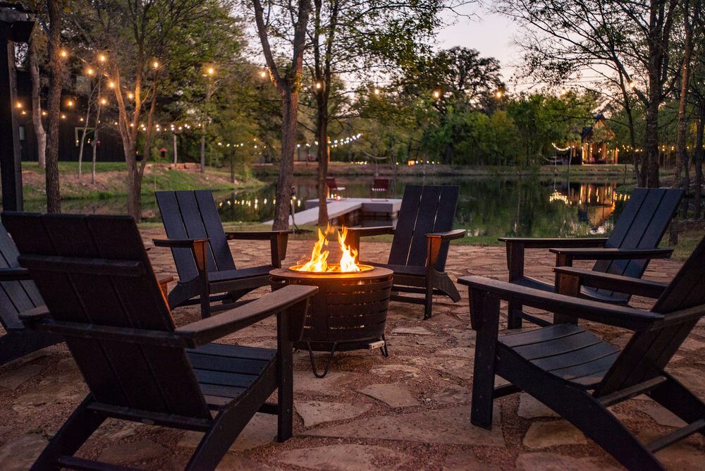 Šest crnih stolica poredanih oko okruglog stola s vatrom u sredini. Iznad stola su obješene žarulje, a u pozadini se vidi jezero i drveće