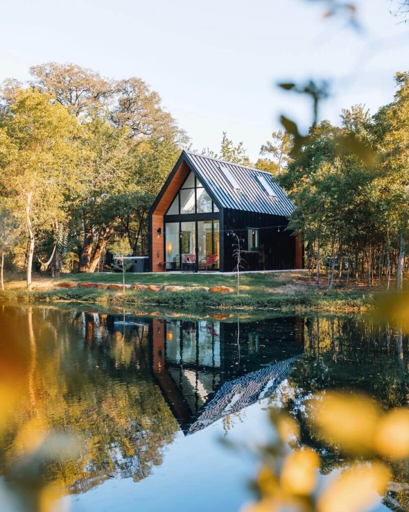 Mala moderna crna kuća s velikim staklenim prozorima i kosim krovom, smještena uz mirno jezero koje zrcali kuću i okolni šumoviti krajolik