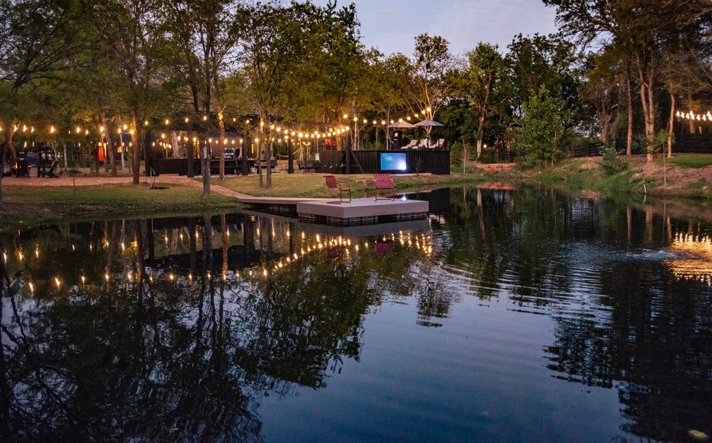 Jezero okruženo drvećem, s platformom na vodi na kojoj se nalaze dvije stolice. Iznad jezera su obješene žarulje, stvarajući ugodnu atmosferu