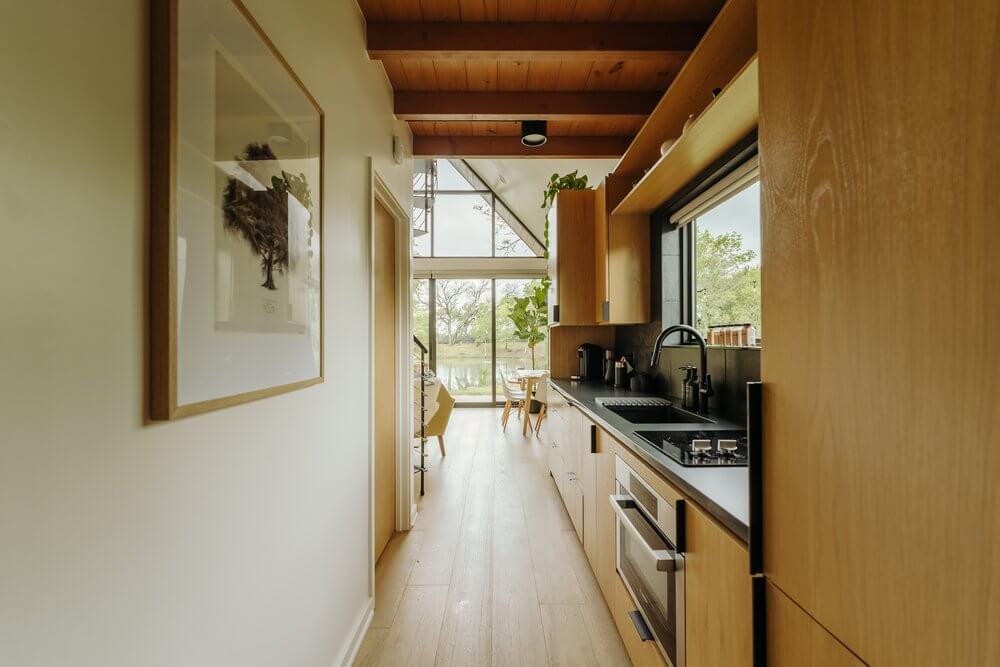 Hodnik untar kućice s kuhinjom u pozadini. Hodnik ima svijetle zidove i drveni pod, a kuhinja je opremljena modernim, minimalističkim elementima