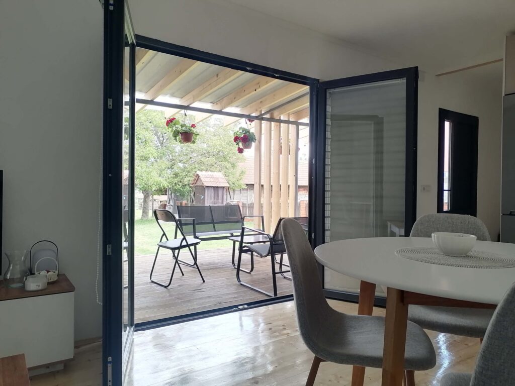 Otvorena staklena vrata vode na terasu s namještajem, okruženu zelenilom i drvećem