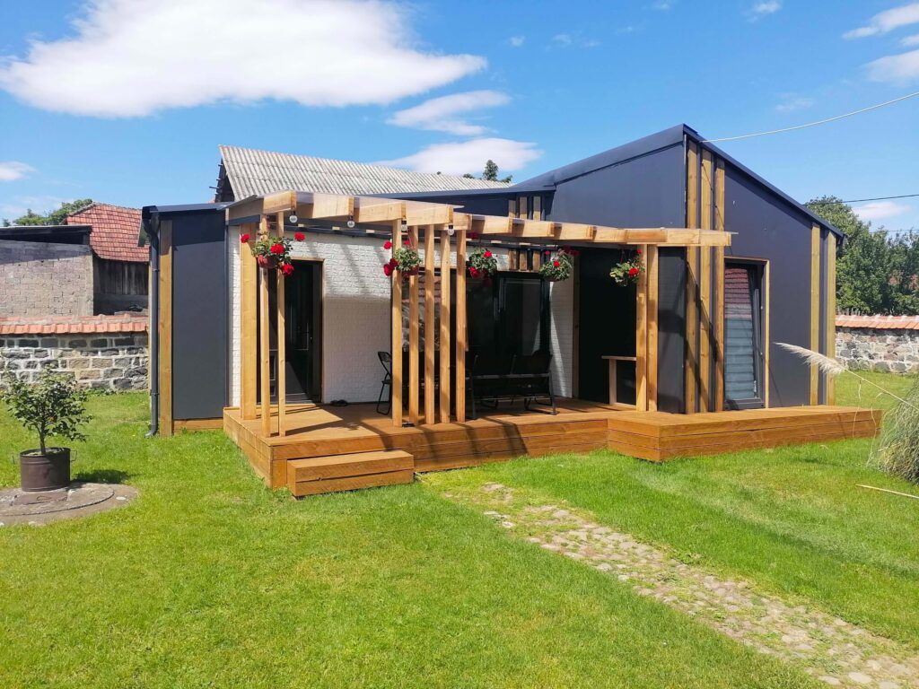 Moderna, tamna modularna kućica sa drvenom pergolom i terasom u zelenom dvorištu