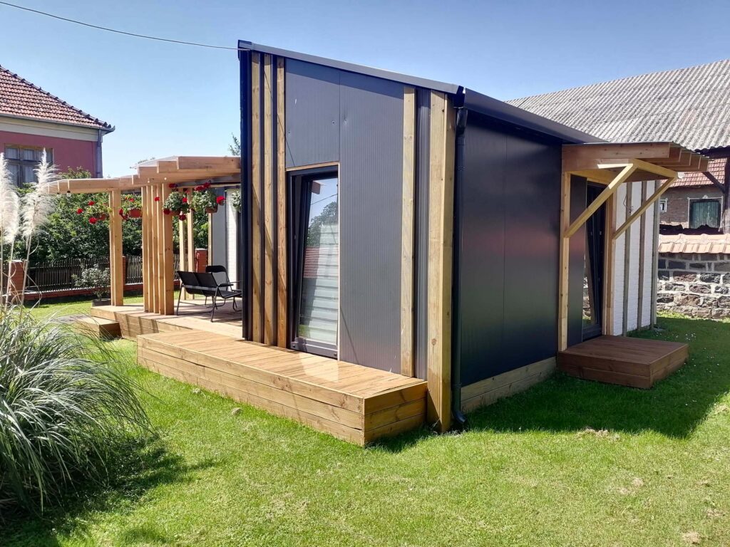 Moderna modularna kuća s drvenom terasom, pergolom i vanjskim namještajem, okružena zelenim dvorištem