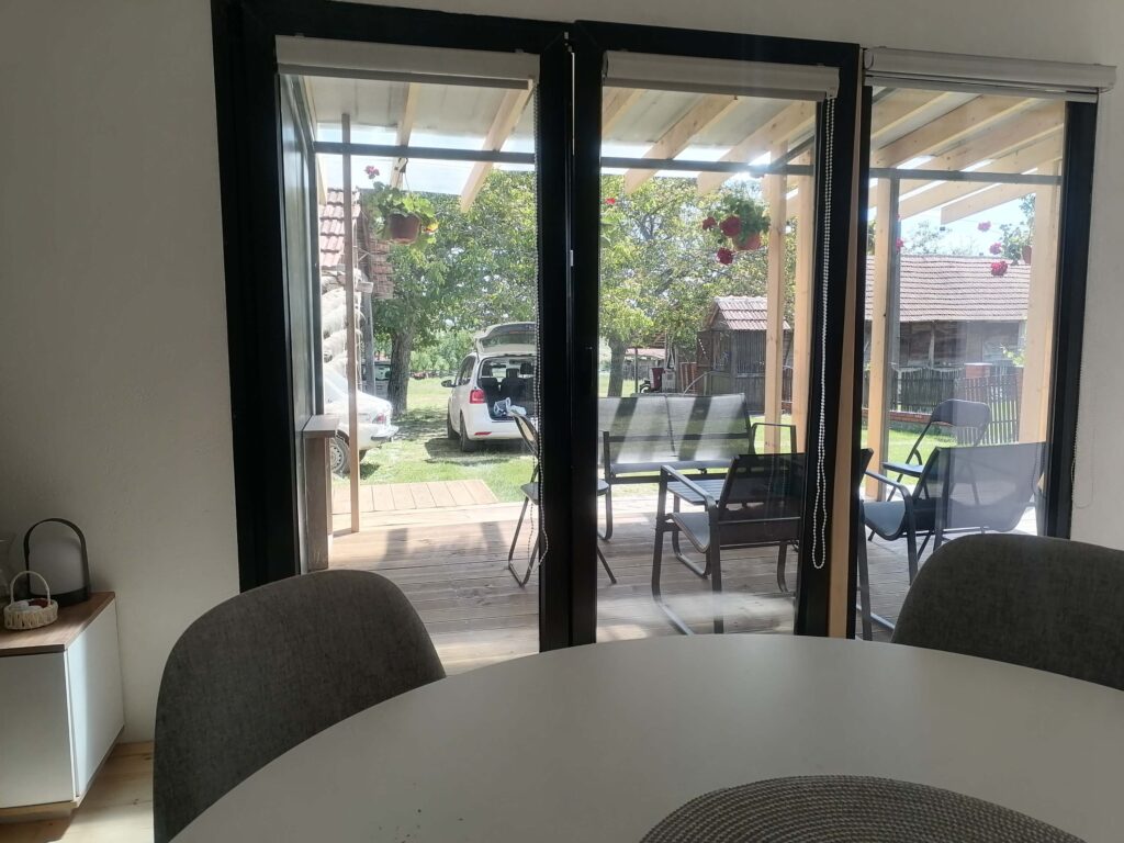 Panoramska staklena vrata modularne kućice pružaju pogled na terasu s namještajem, dvorište s automobilom i zelenilom
