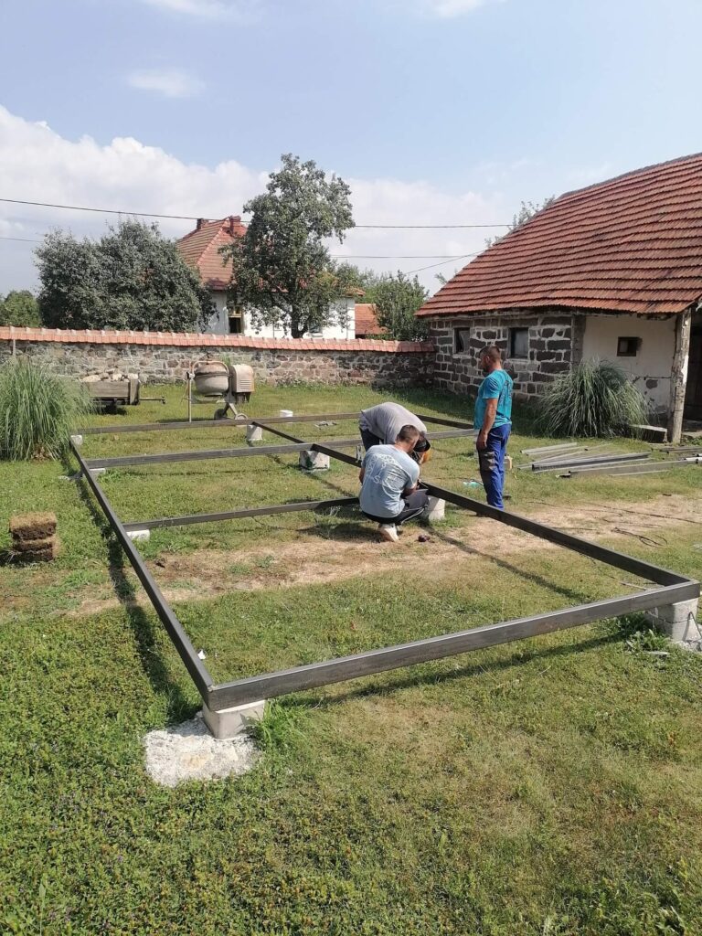 Tri radnika sastavljaju metalnu konstrukciju za temelj modularne kućice na travnatom terenu