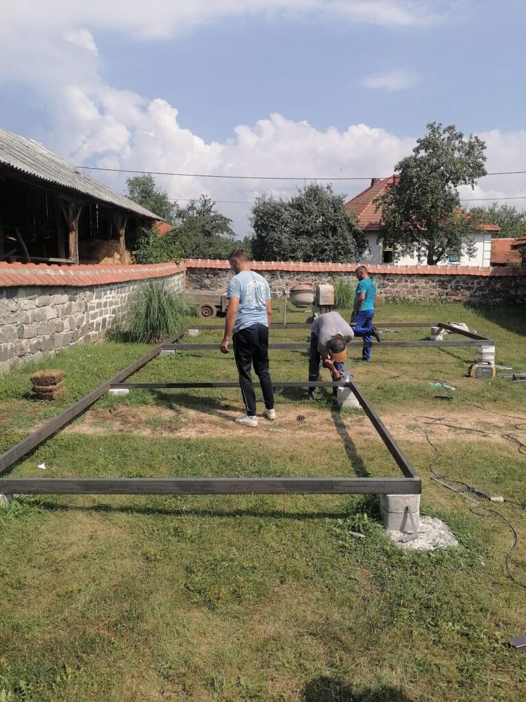 Tri radnika sastavljaju metalnu konstrukciju za temelj modularne kućice na travnatom terenu