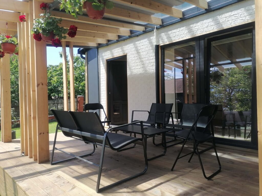 Moderna terasa s drvenim podom i pergolom s visećim cvjetnim loncima. Na terasi se nalazi crni metalni stol sa četiri stolice, idealan za sjedenje na otvorenom