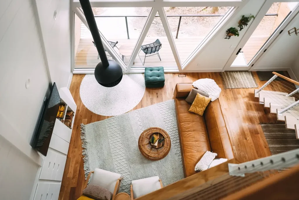 Unutrašnjost A-frame vikendice, pogled odozgo na dnevni boravak s kožnim kaučem, modernim kaminom i visokim prozorima