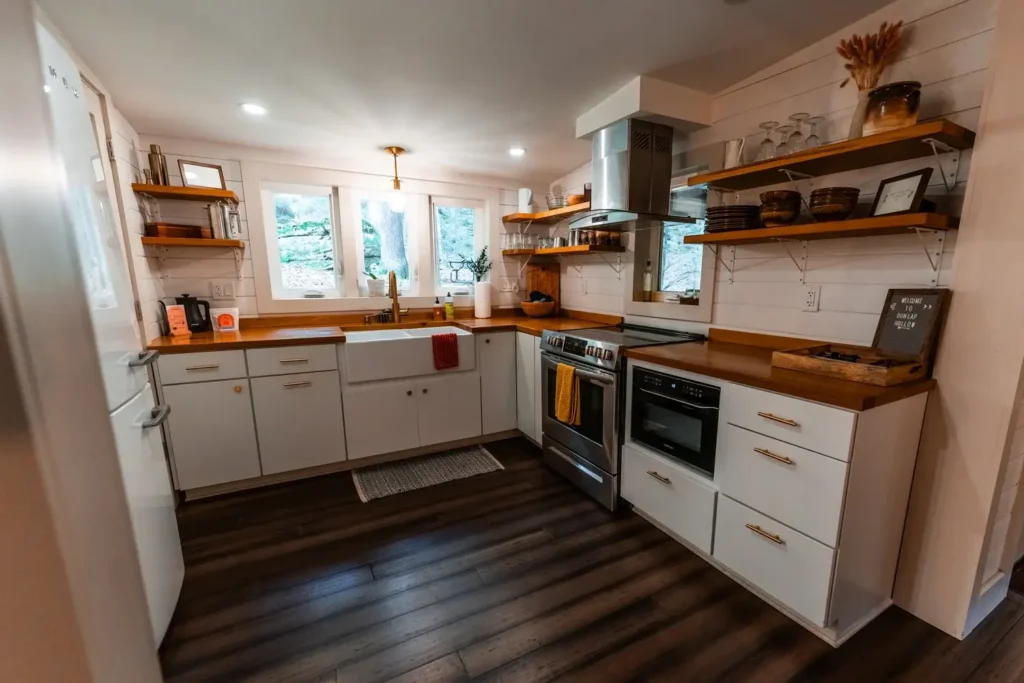 Modernistička kuhinja u kolibi s bijelim ormarićima, drvenim pultovima, otvorenim policama i sudoperom