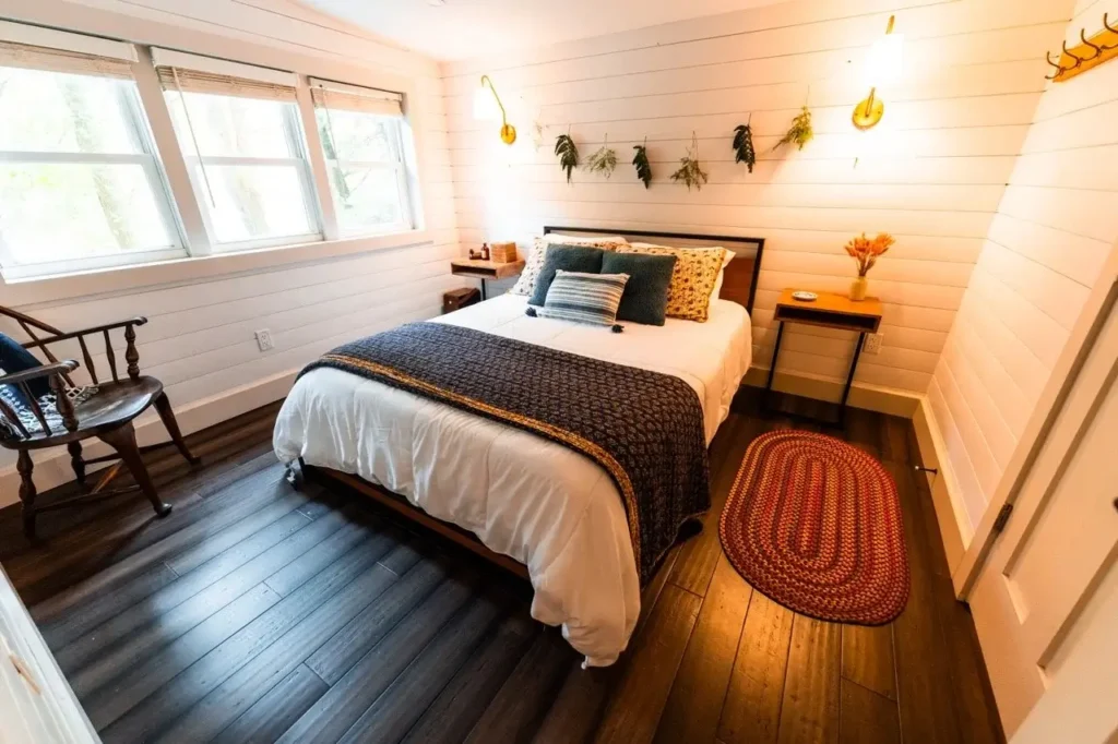 Spavaća soba s bračnim krevetom, drvenim podom, bijelim zidovima i biljkama koje vise iznad uzglavlja