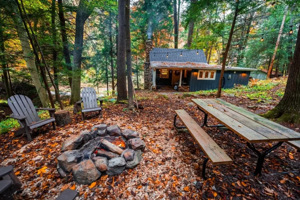 Rustikalna koliba u šumi s logorskom vatrom, drvenim stolom za piknik i stolicama, okružena drvećem i lišćem