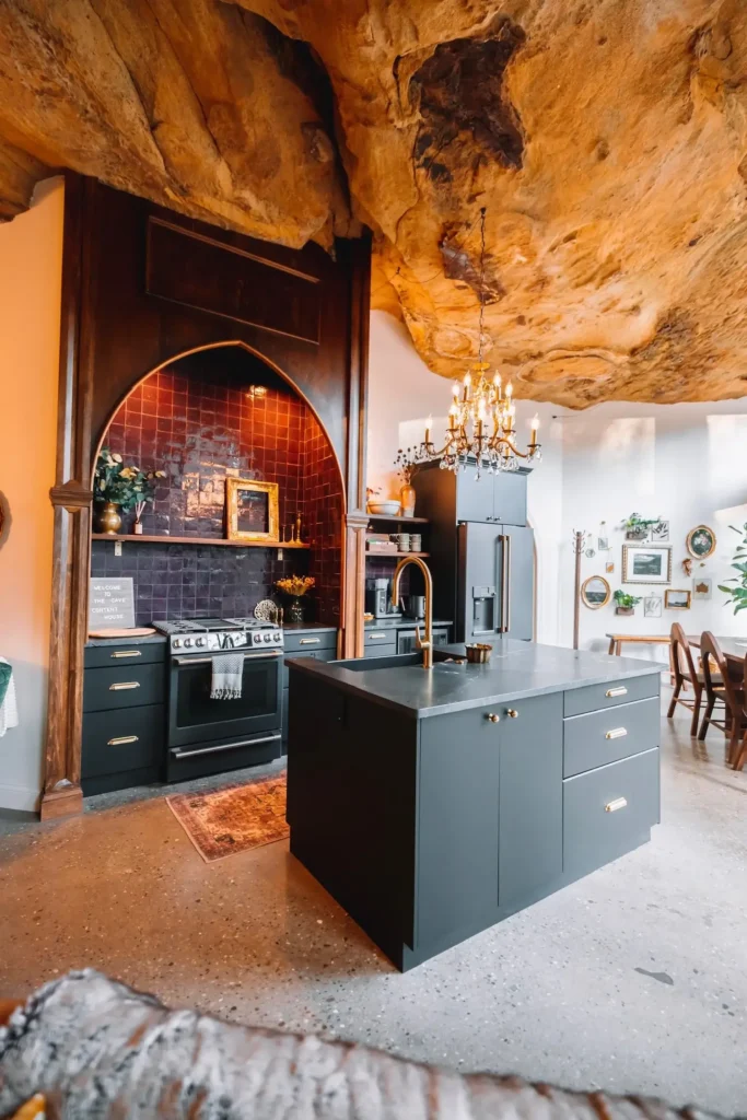 Moderno opremljena kuhinja s tamnim ormarićima, kuhinjskim otokom, zlatnim slavinama i kamenim stropom