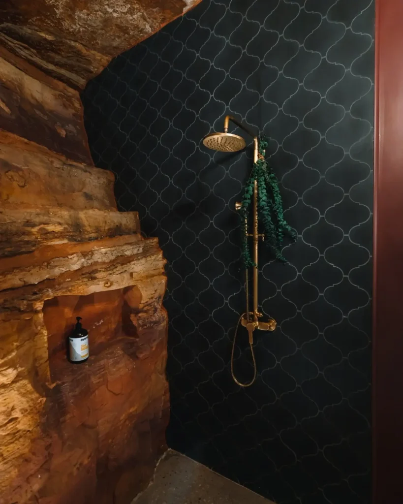 Rustikalna tuš kabina s tamnim mozaik pločicama i zlatnom slavinom. Lijevo je zid nalik stijeni s policom