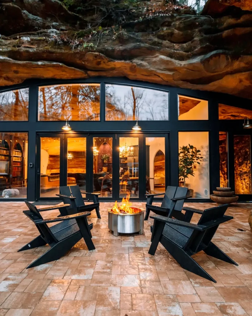 Rustikalna terasa s četiri crne stolice oko ložišta. Ispred je vikendica od prirodne stijene s velikim prozorima