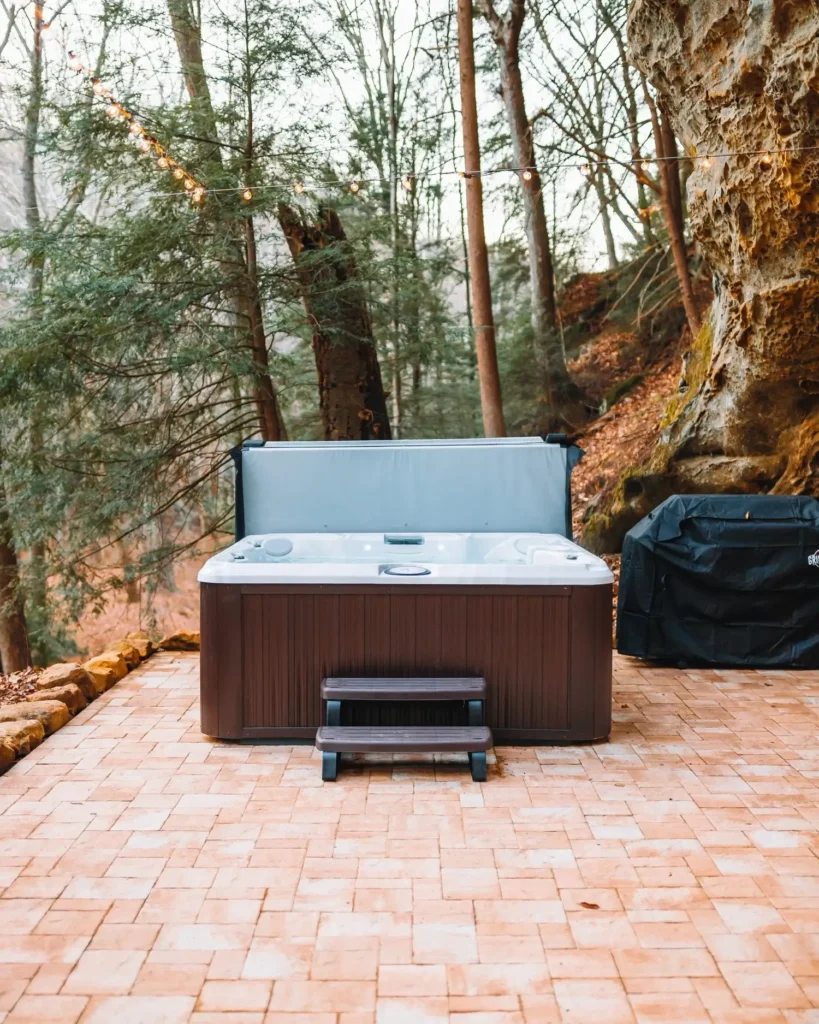 Vruća kupelj na terasi s pločicama, okružena drvećem i stijenama. U pozadini je šuma