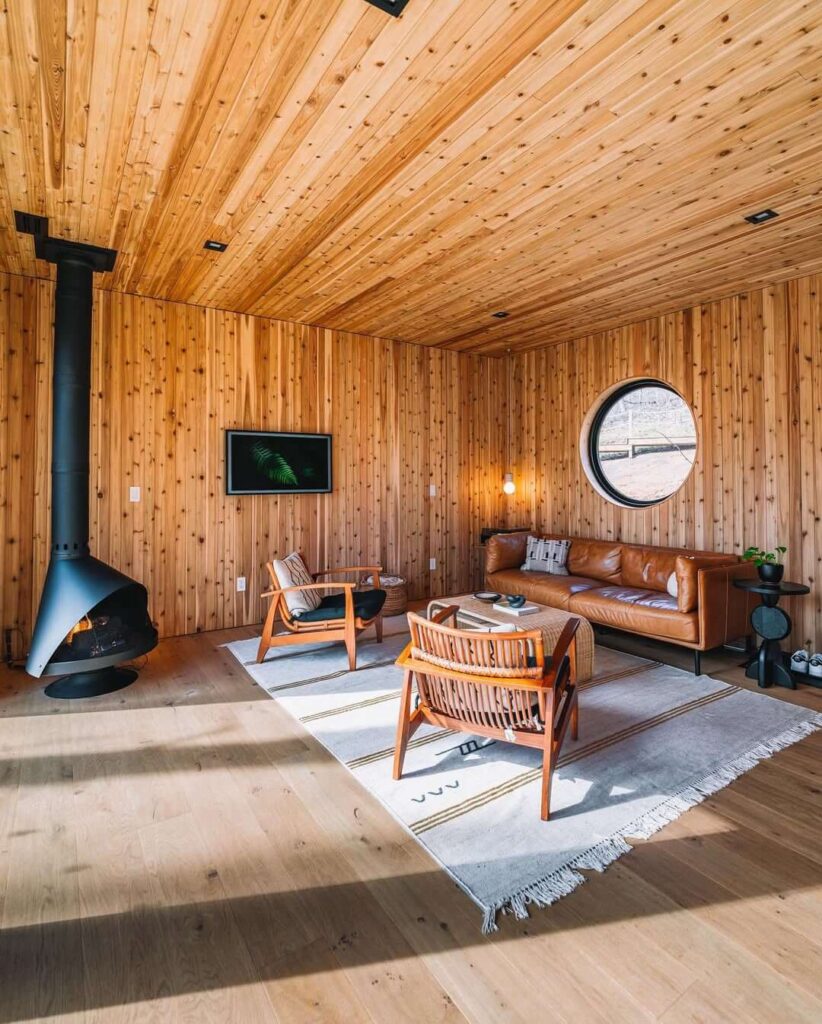 Dnevni boravak u bungalovu s drvenim zidovima i stropom, visećim kaminom i okruglim prozorom
