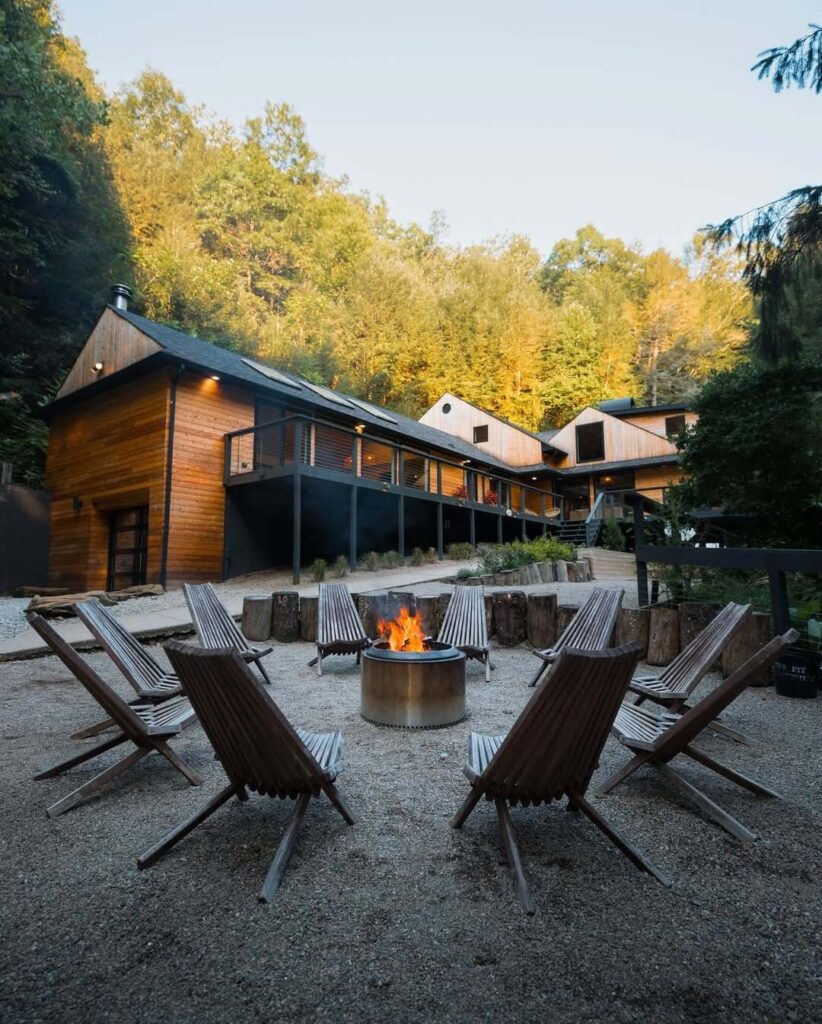 Vanjsko ognjište okruženo drvenim stolicama, s modernom drvenom kućom i šumom u pozadini