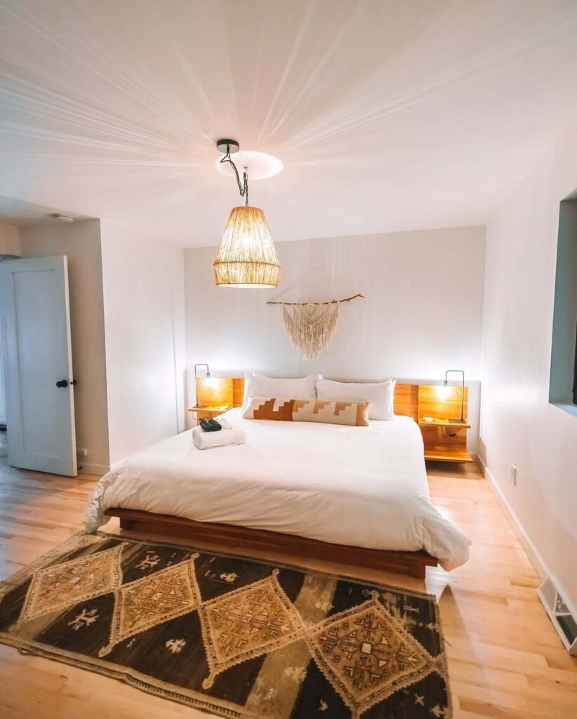 Svijetla spavaća soba s drvenim krevetom, pletenim lusterom, boho tepihom i bijelim zidovima