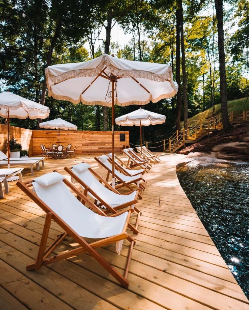 Drvena terasa uz bazen s redom bijelih ležaljki i suncobranima, okružena šumom