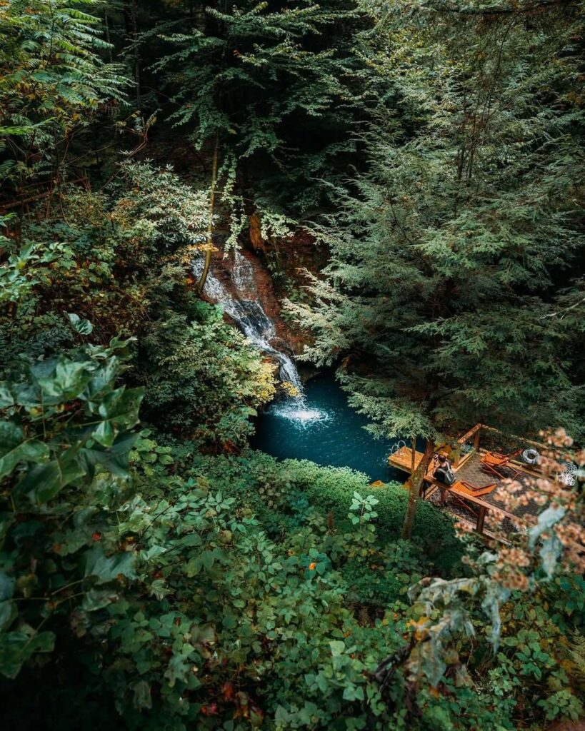 Pogled iz zraka na mali vodopad koji se ulijeva u bazen, okružen gustom šumom i drvenom platformom