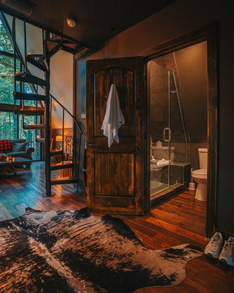 Moderna kupaonica s tamnim pločicama i staklenom tuš kabinom uz rustikalna drvena vrata u A-frame kućici