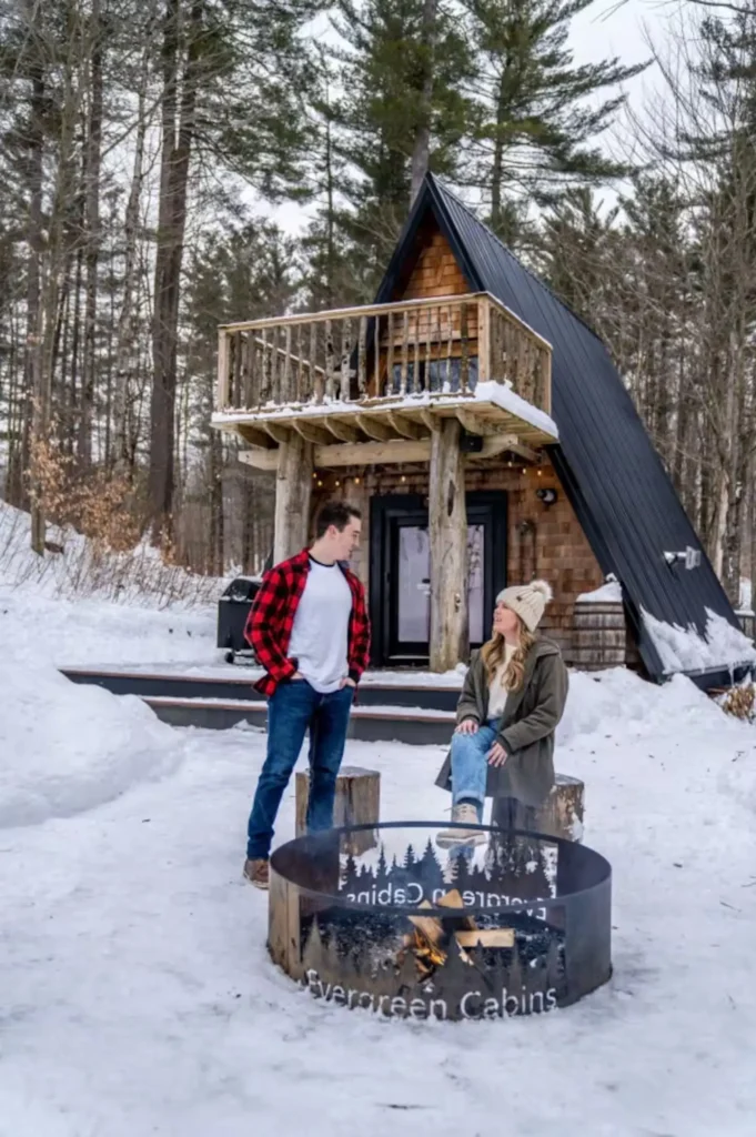 Mladi par stoji pored ognjišta ispred drvene A-frame kućice na snijegu u šumi