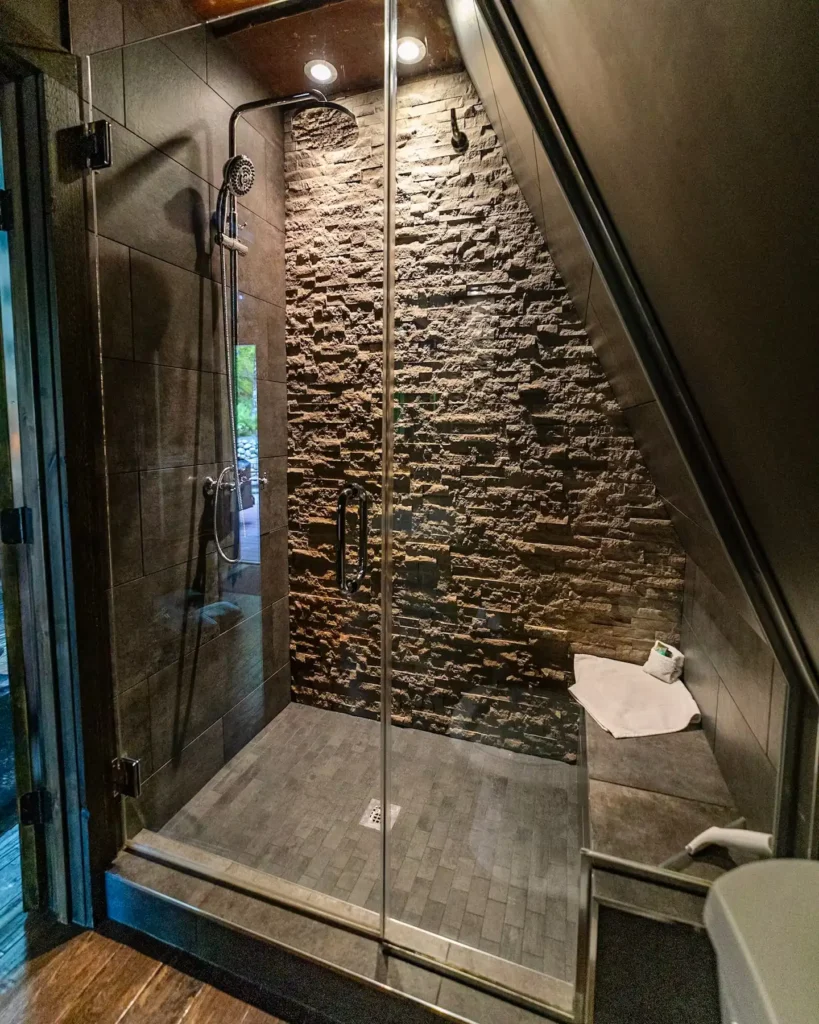 Luksuzna tuš kabina sa zidom od prirodnog kamena i tamnim podnim pločicama u modernoj A-frame kolibi