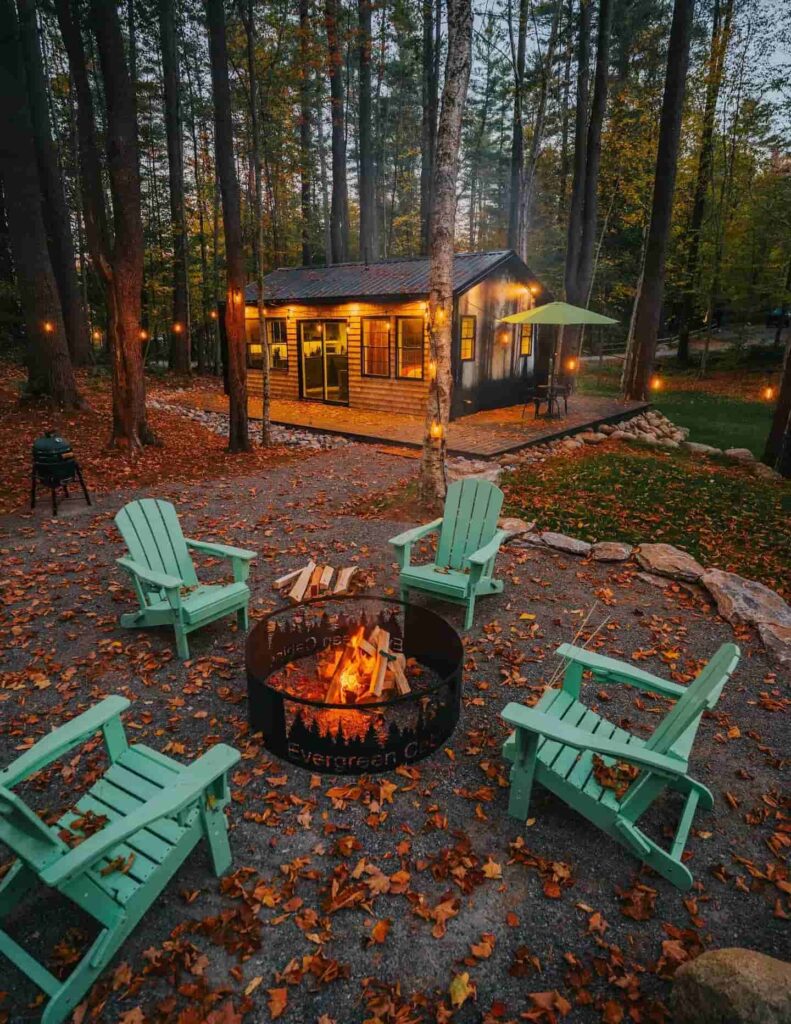 Osvijetljena drvena koliba u šumi s tirkiznim stolcima oko ognjišta prekrivenog jesenskim lišćem