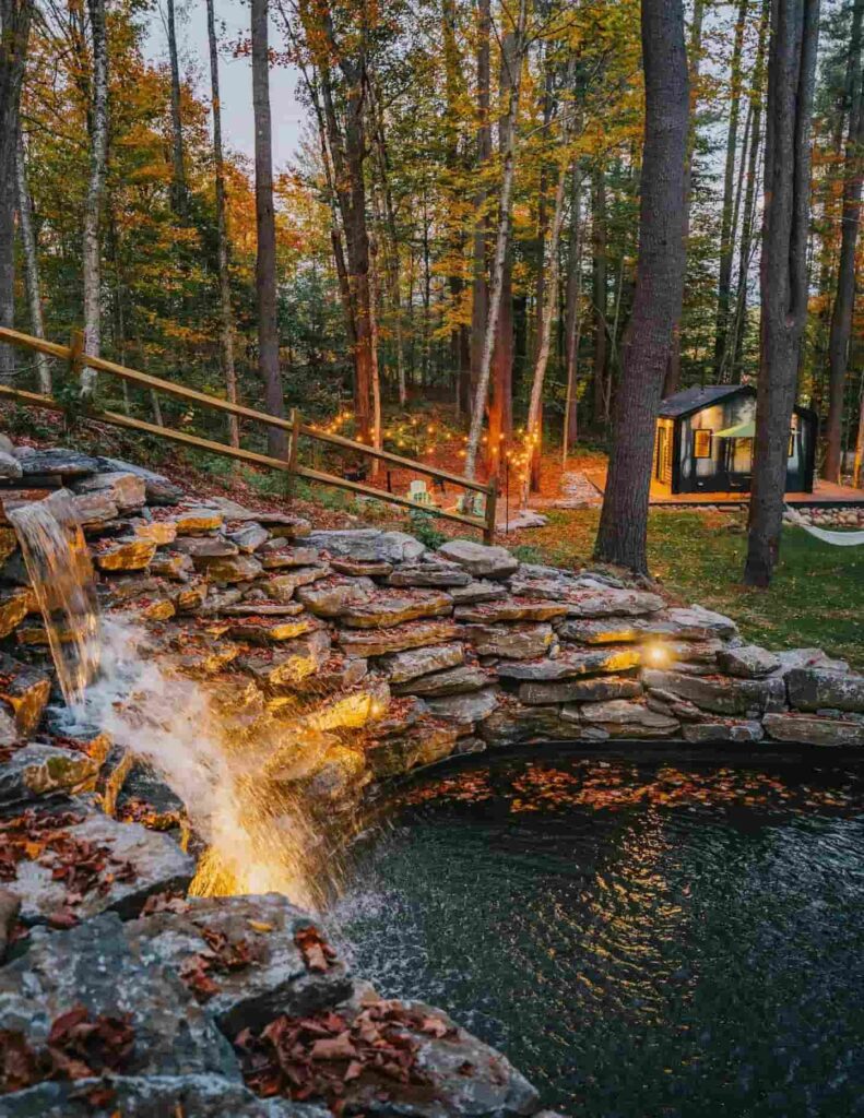 Umjetni kameni vodopad i bazen u jesenskoj šumi s osvijetljenom drvenom kolibom u pozadini