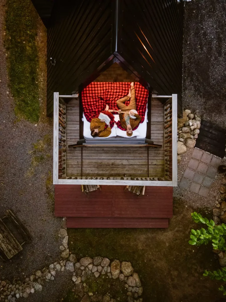 Pogled iz zraka na par koji leži na krevetu izvučenom na terasu A-frame kućice usred šume