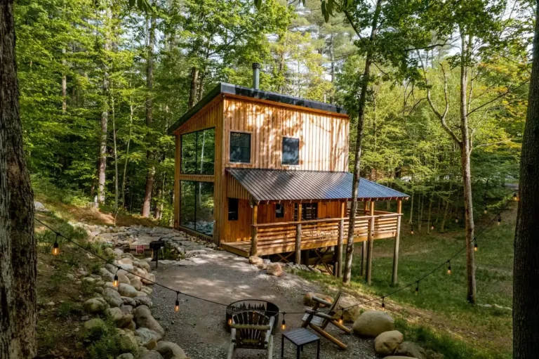 Moderna drvena koliba u šumi s velikim prozorima, terasom i vanjskim ognjištem okružena drvećem