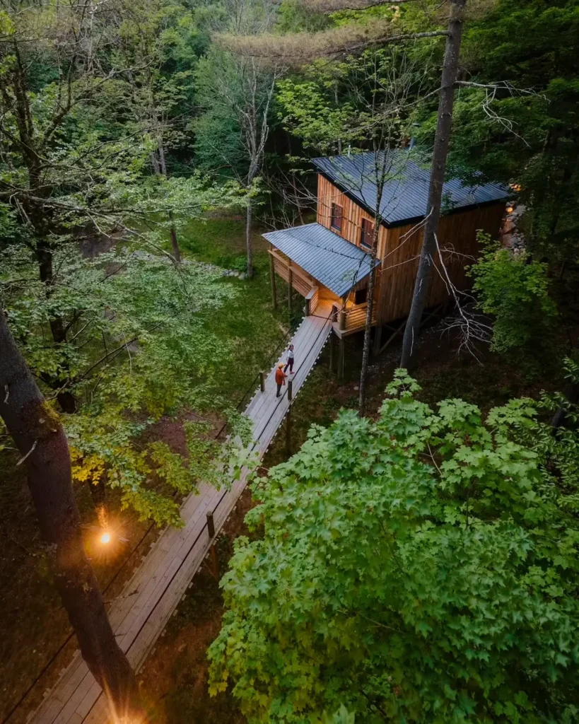 Pogled iz zraka na osvijetljenu drvenu kućicu u šumi do koje vodi dugački drveni viseći most