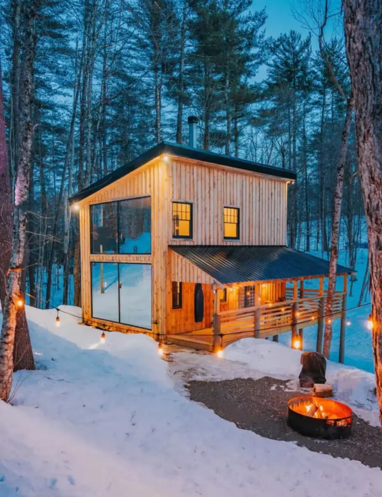Osvijetljena drvena koliba u snježnoj šumi s upaljenim ognjištem i dekorativnim lampicama navečer