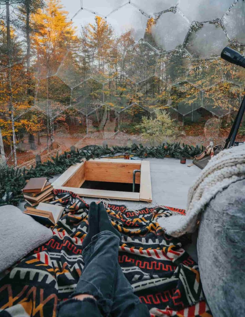 Unutrašnjost prozirne kupole s pogledom na jesensku šumu, knjigama, teleskopom i toplom dekom za opuštanje