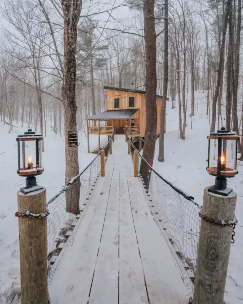 Drveni viseći most s fenjerima vodi do osvijetljene kolibe u snježnoj zimskoj šumi