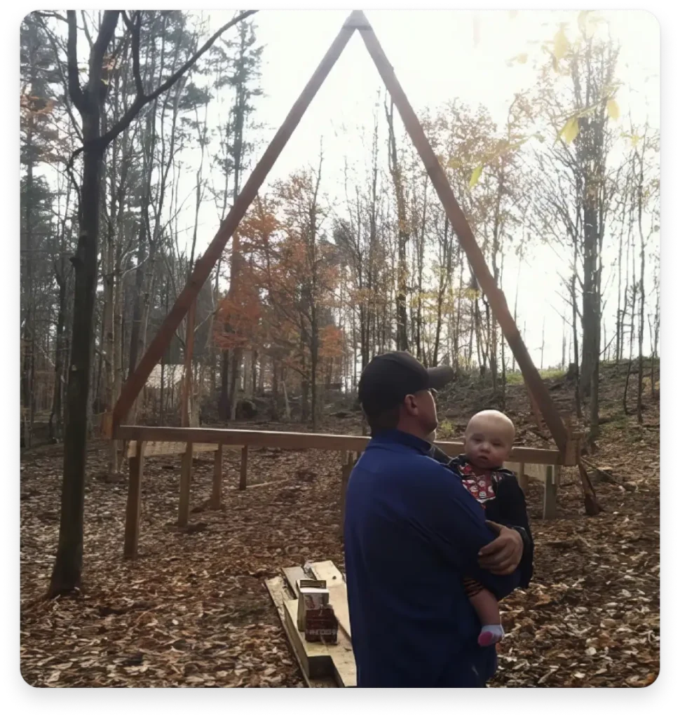 Muškarac drži bebu ispred drvene konstrukcije u obliku trokuta usred šume.