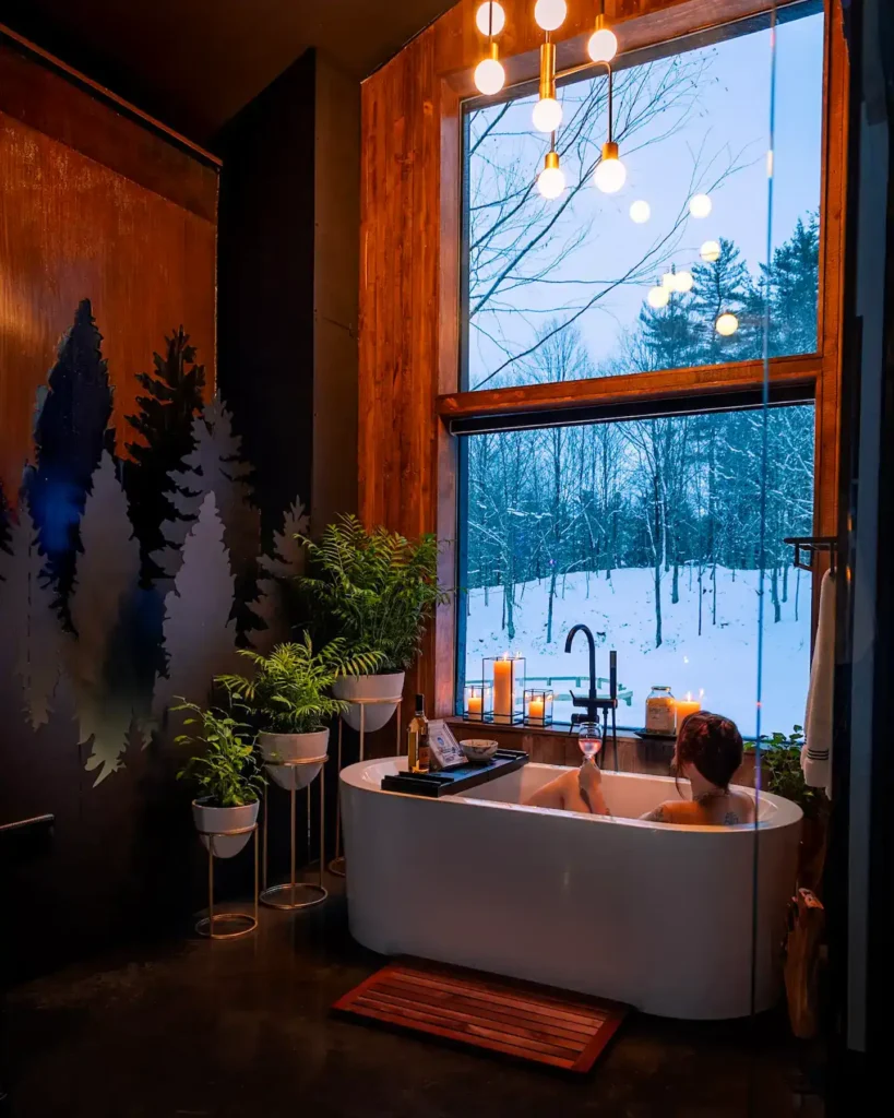 Žena se opušta u kadi uz svijeće i pogled kroz veliki prozor na snježnu šumu